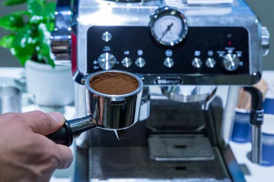 Hand holding ground coffee in a portafilter in front of a De'Longhi La Specialista espresso machine