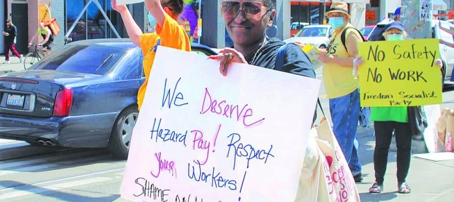 A woman holds a sign that reads: "We Deserve Hazard Pay! Respect Your Workers! Shame on you Kroger!" In the background another sign reads "No safety No work - Freedom Socialist Party"