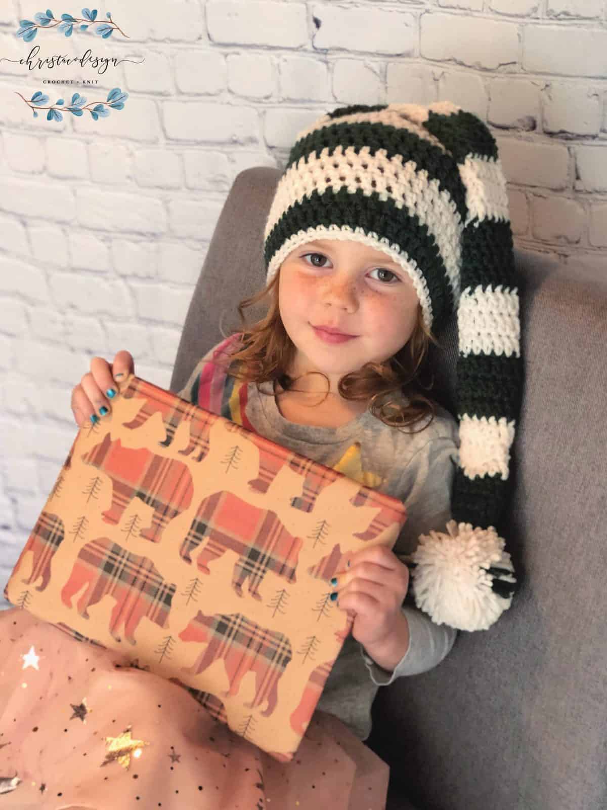 picture of girl with long striped stocking hat on in green and white holding a wrapped Christmas gift in bear paper