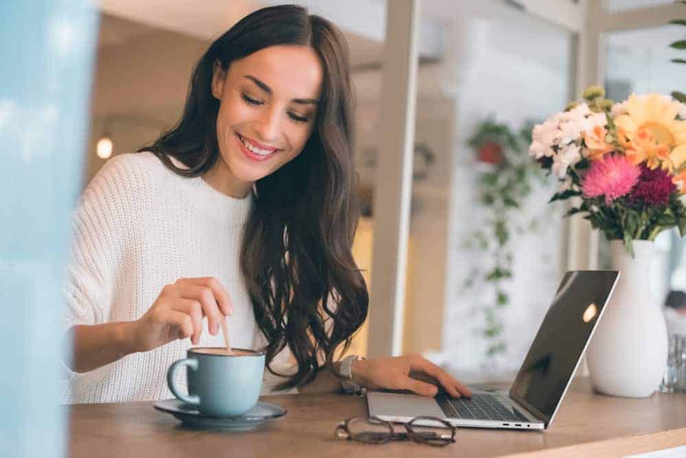 smiling woman stirring mug of coffee while holding her other hand on a laptop with colorful flowers nearby