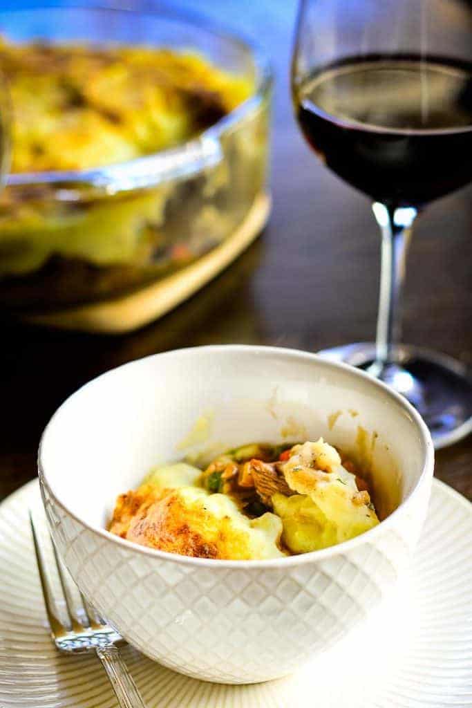 Vertical shot of a single serving of Classic Lamb Shepherd's Pie in a white bowl with a glass of red wine and the serving dish in the background. Text Overlay: Lamb Shepherd's pie