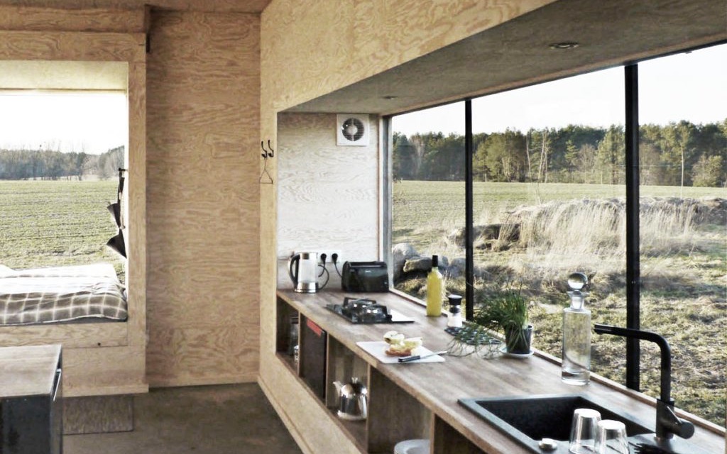 Das Bild zeigt ein Tiny House von innen. Im Tiny House sieht man eine Küche mit einem großen Fenster und einem Schlafplatz.