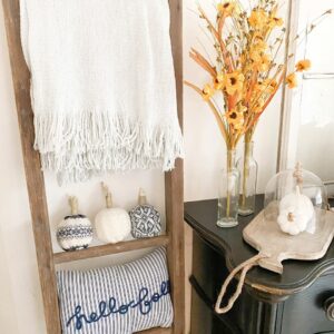 Photo shows the 3 completed DIY sweater pumpkins sitting on an antique ladder underneath a grey throw blanket and above a blue and white striped pillow that says "hello fall" It also shows a black antique looking dresser with some yellow paper flowers in some clear bottle vases, a wood tray with 2 ceramic white pumpkins underneath a glass cloche.