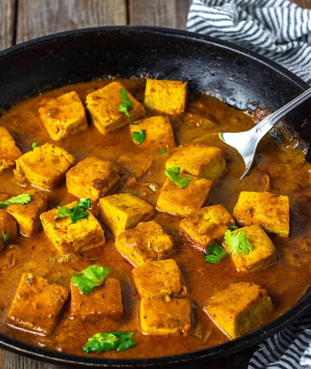 Tofu tikka masala straight on view in a black skillet with lots of curry sauce, garnished with cilantro leaves on a wooden background with a silver spoon