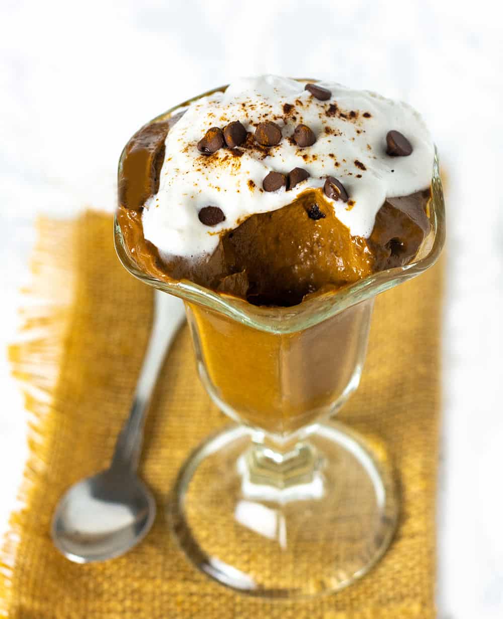 Carob Mousse in a glass jar topped with coconut cream sprinkled with carob chips. One scoop of mousse removed glass on a burlap napkin on a white marble background