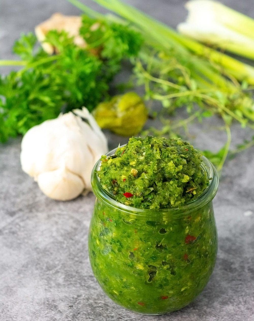 green seasoning recipe in a glass jar on a grey marble background with celery, garlic, pepper, ginger in the background