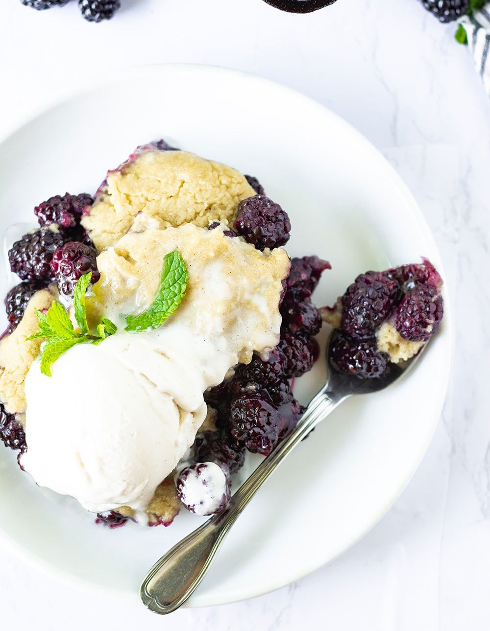 overlay vegan blackberry cobbler on a white plate with a scoop of vegan vanilla ice cream and mint leaf garnish with a silver spoon with cobbler on a white background