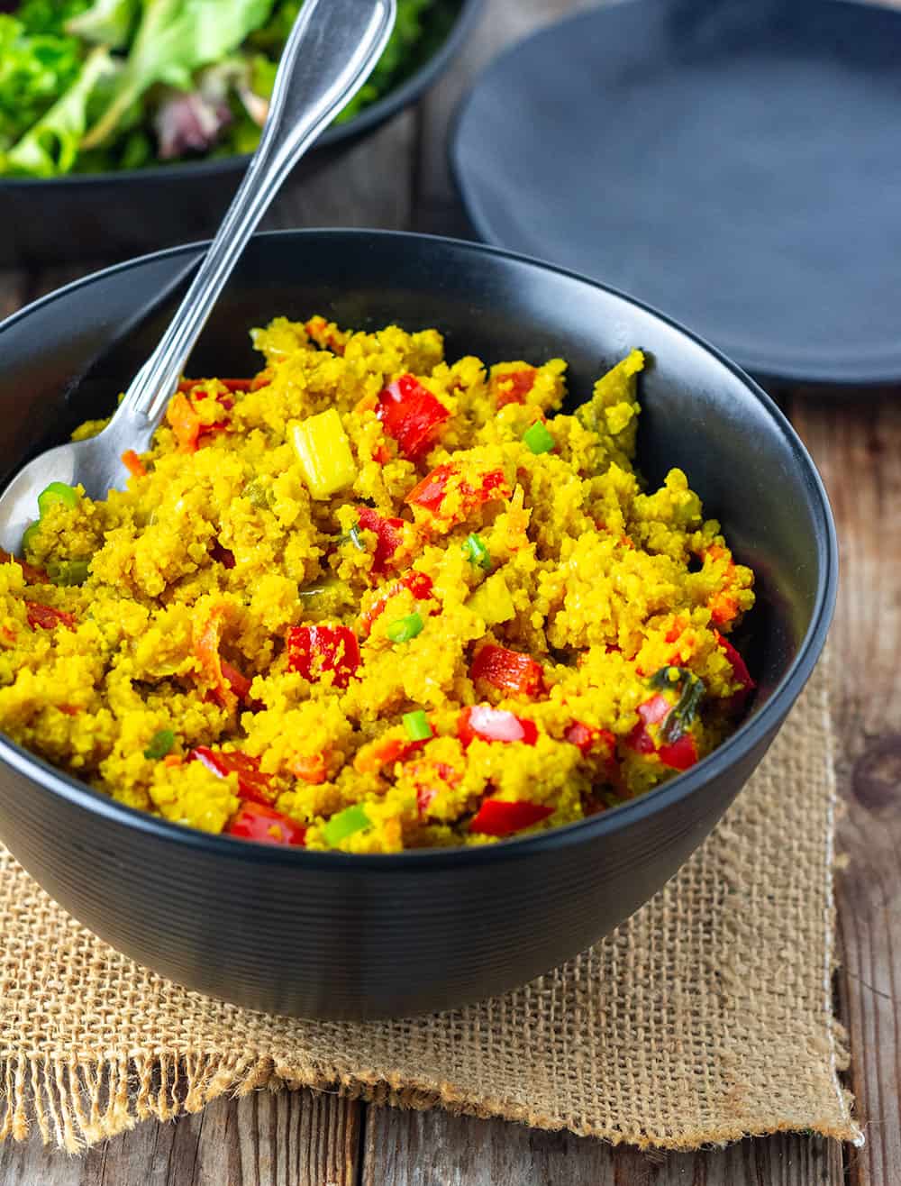 Cauliflower rice curry with red bell pepper, chopped green onion, carrot strips in a black bowl, silver spoon on a brown burlap napkin on a wooden background