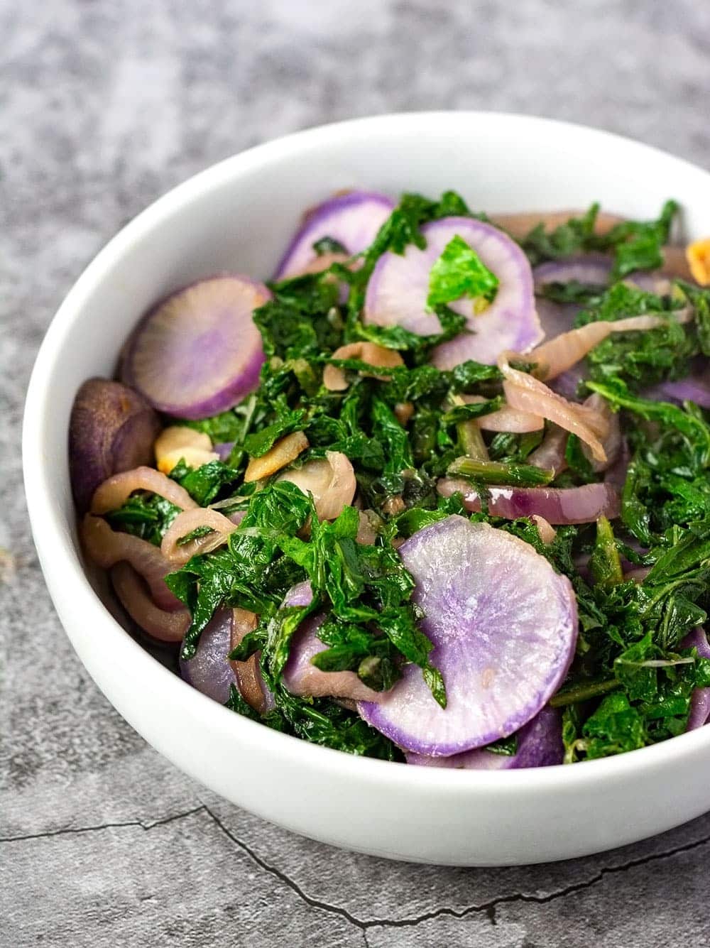 Green sauteed radish leaves and slices of purple sauteed radishes in a white bowl over a grey marbled background