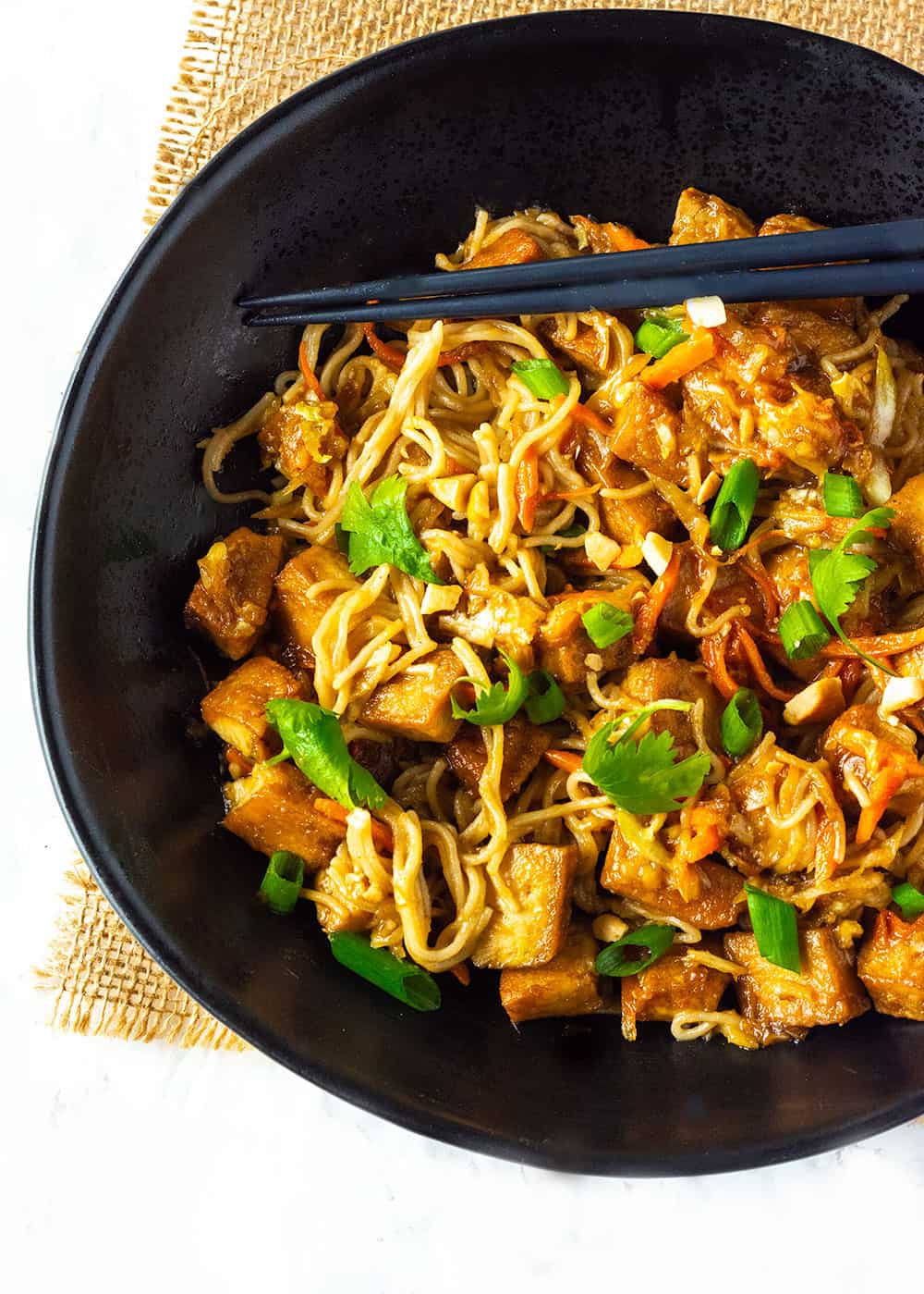 Peanut Noodles With Tofu in a black bowl, with black chop sticks and garnish with cilantro and peanuts