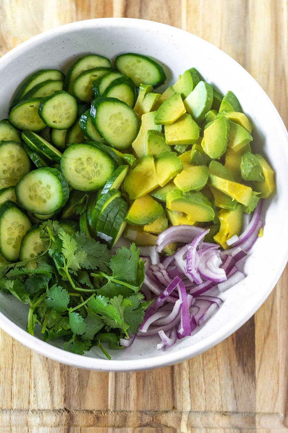 cucumber avocado salad ingredients in a white bowl on a wooden cutting board