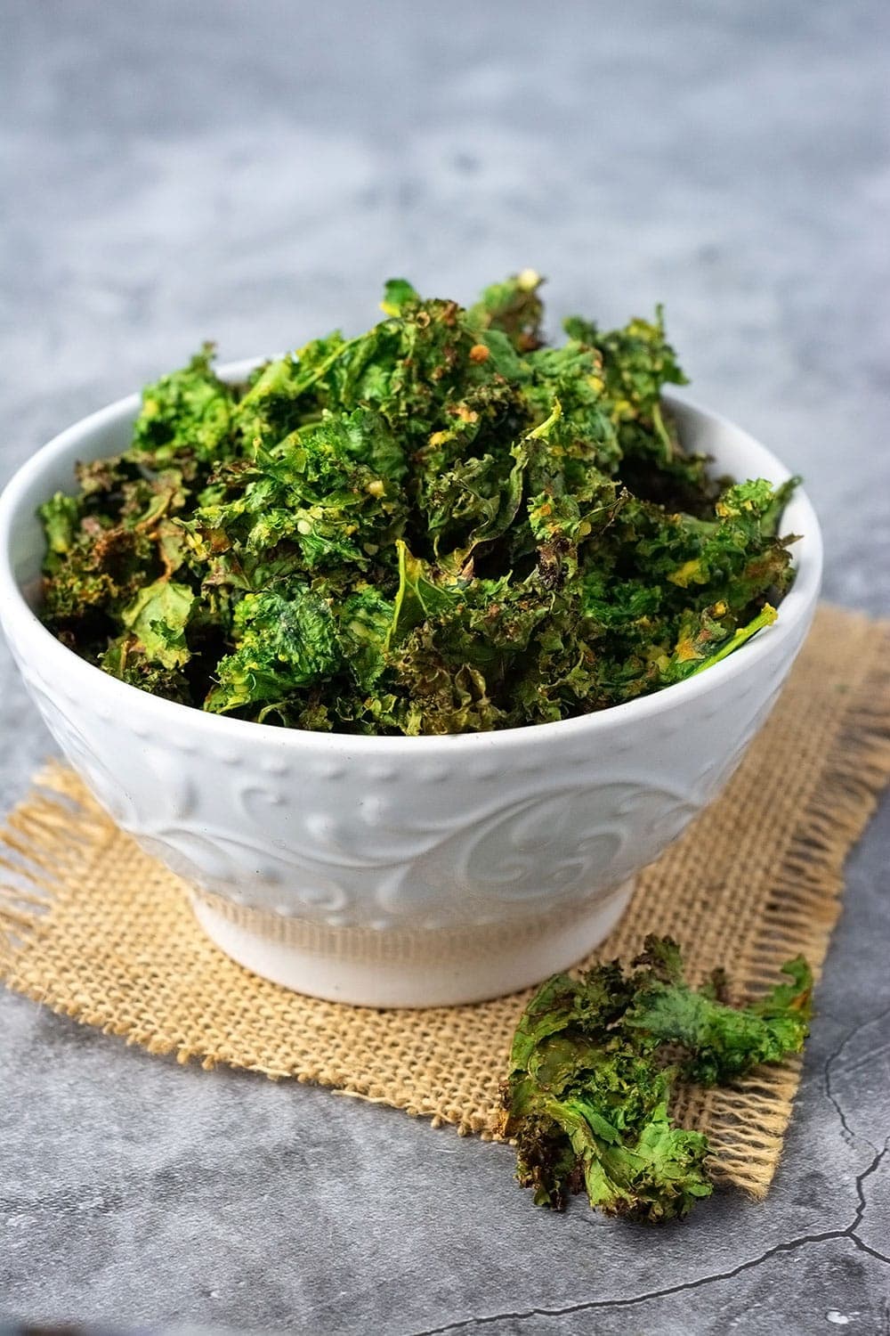 Air fryer kale chips in a white bowl on a burlap napkin on a gray marble background