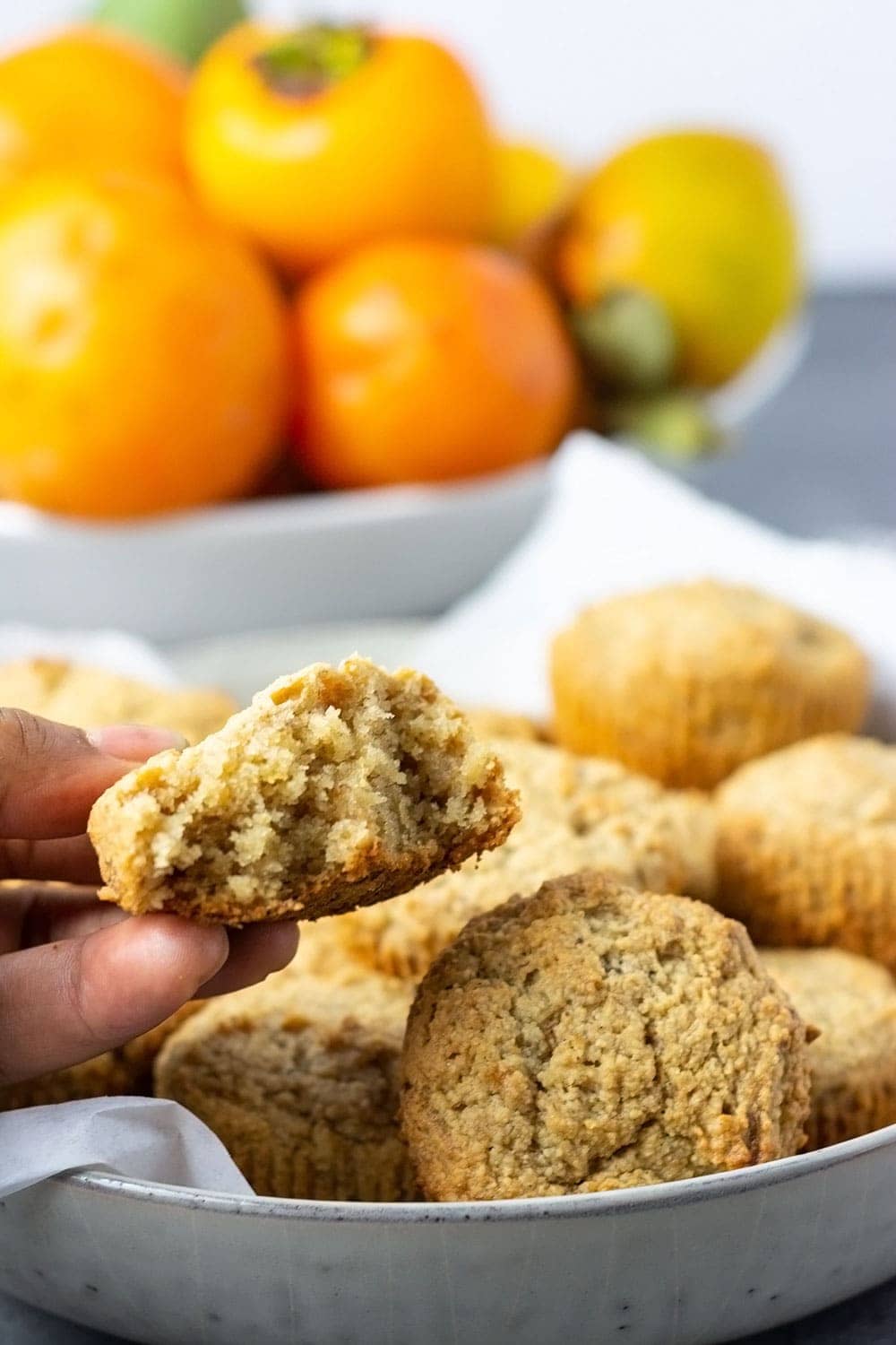 vegan persimmon muffins holding one over a white bowl with more muffins and a bowl of persimmon in a white bowl in the background