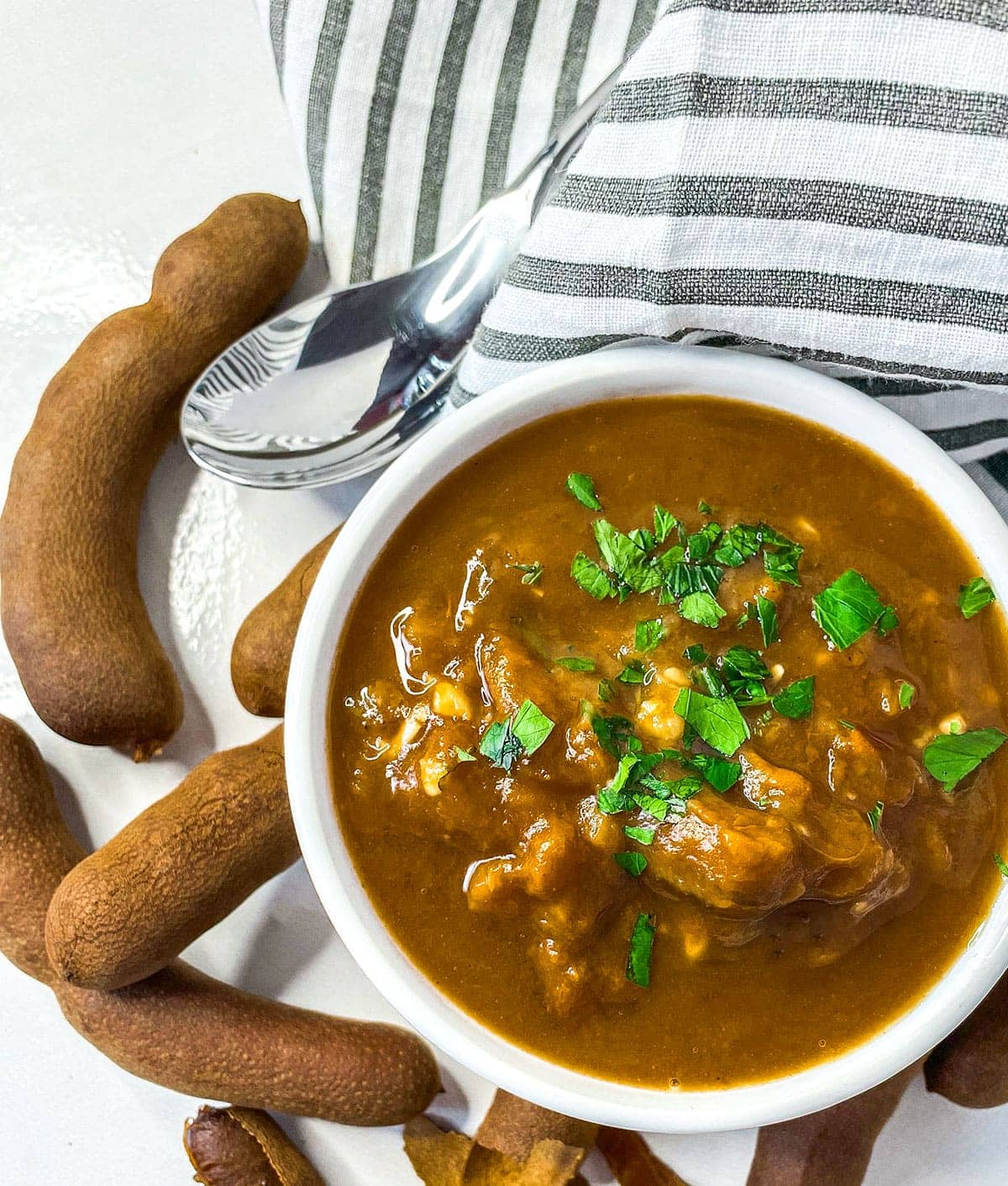 tamarind sauce with tamarind pods surrounding white bowl with a black and white napkin and a silver spoon on a white background