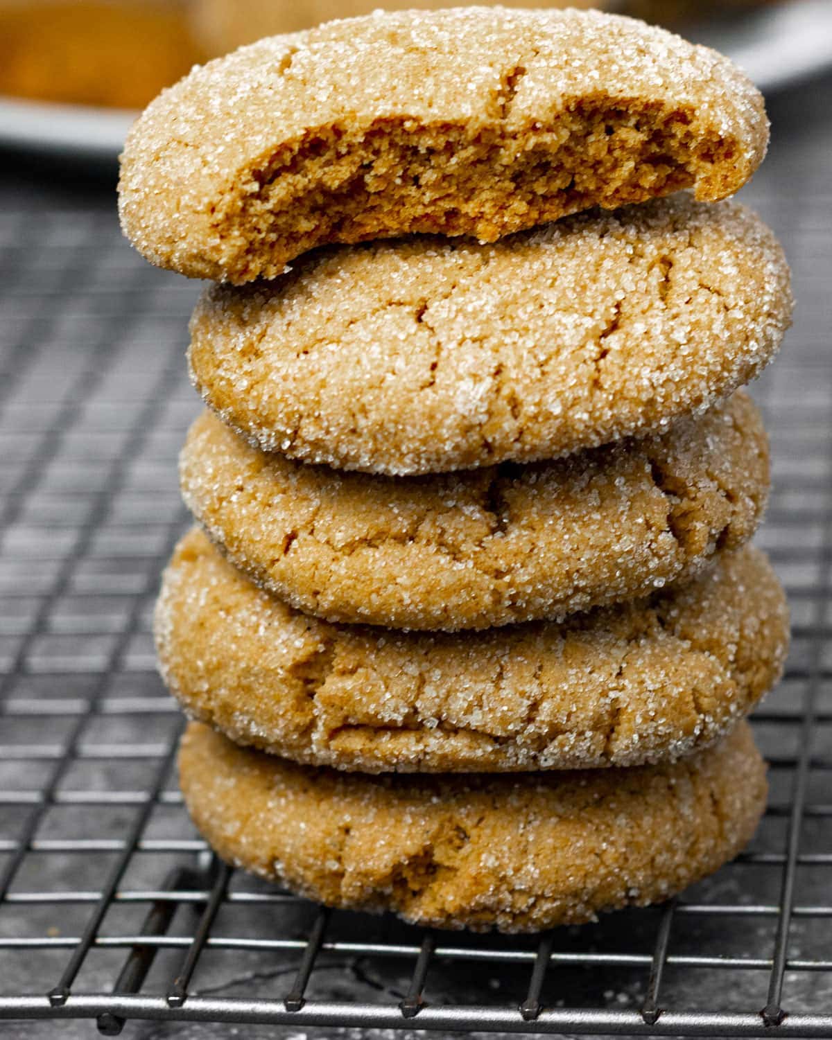 Vegan molasses cookies stacked vertically on a white plate with a plate of cookies in the background. on a burlap napkin on a cooling raf