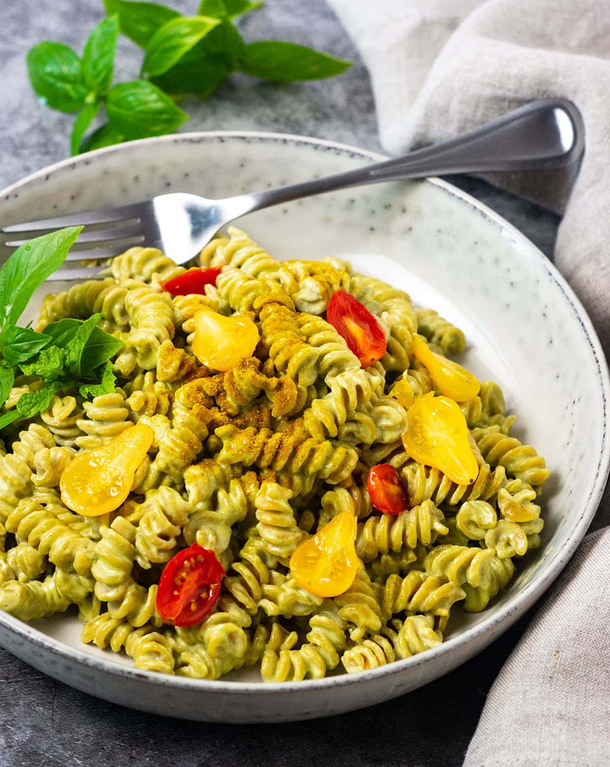 Straight on of creamy vegan pesto pasta in a beige bowl on a grey background with a silver spoon garnished with tomatoes and basil leaves
