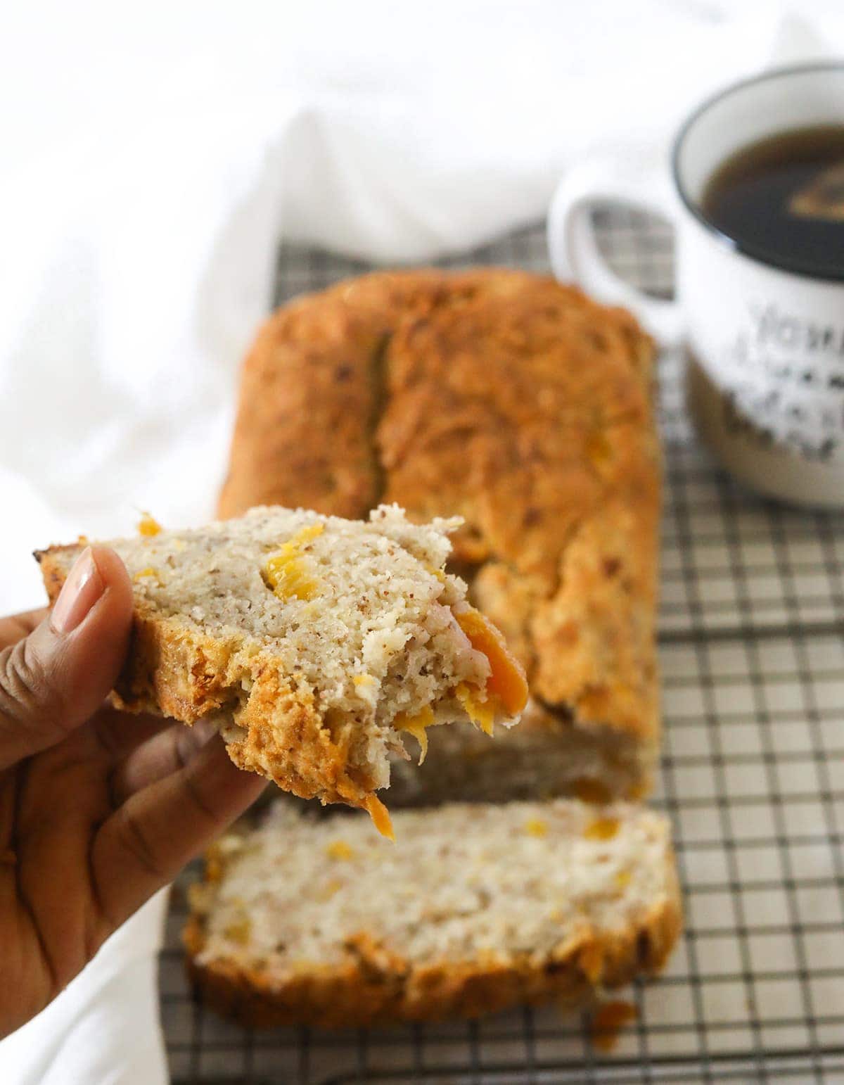 Vegan peach banana bread on a wire rack with two slices, hold one slice in hand on a white background, white napkin and white mug with tea