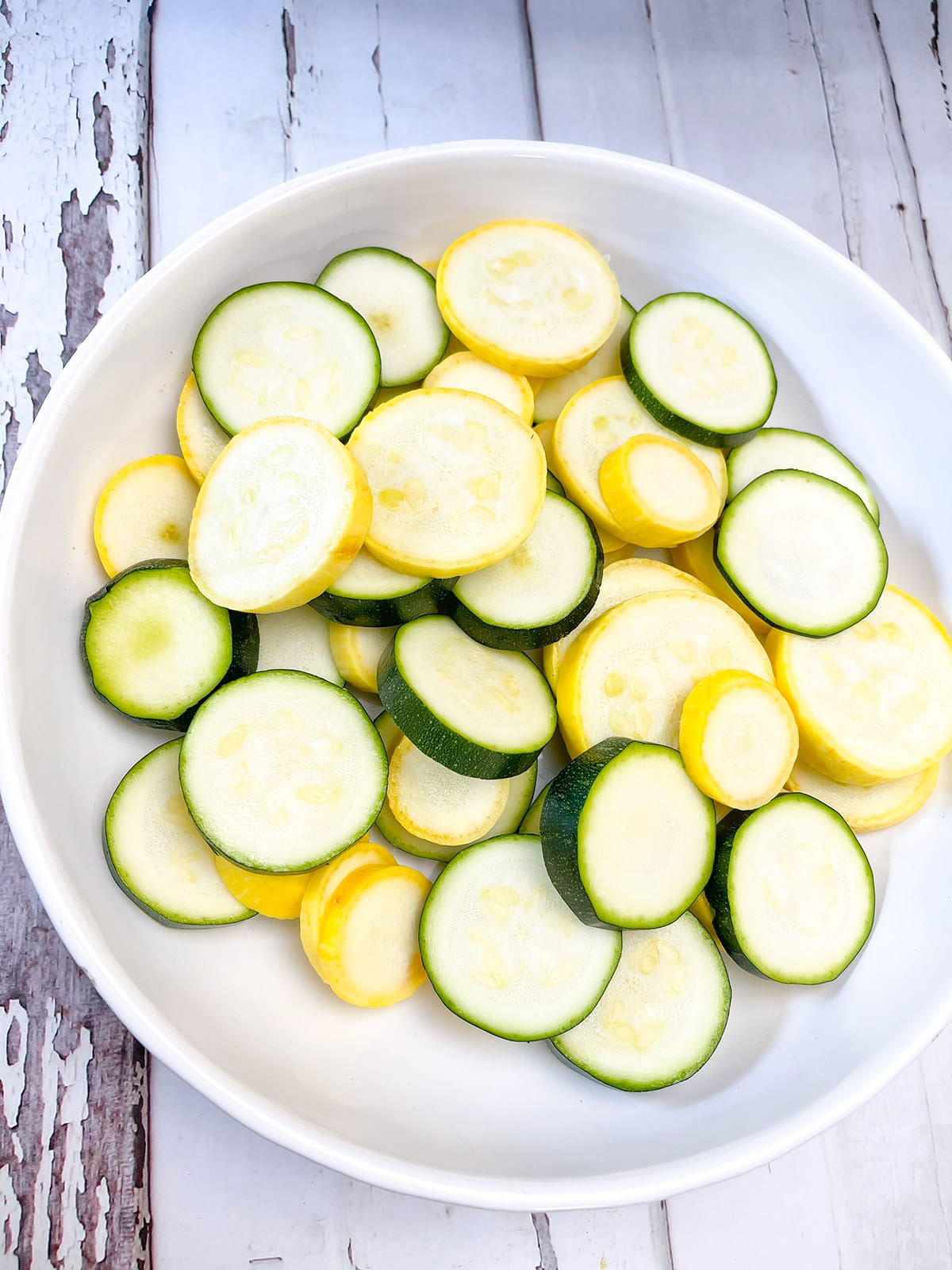 Slices of green and yellow zucchini in a white bowl on a white wooden background