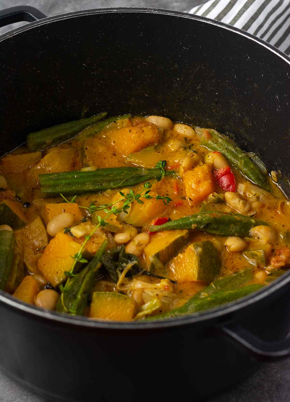 Straight on image butter bean, pumpkin and okra stew in a black pot on a grey background with a black and white stripe.