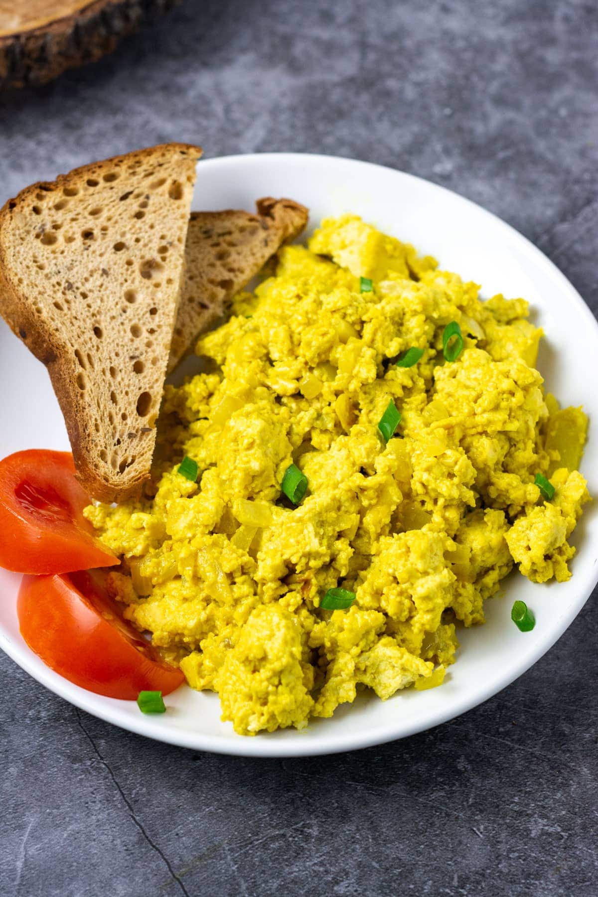 Close up photo easy tofu scramble recipe on a white plate with 2 slices of toasts and tomato wedges on a grey background
