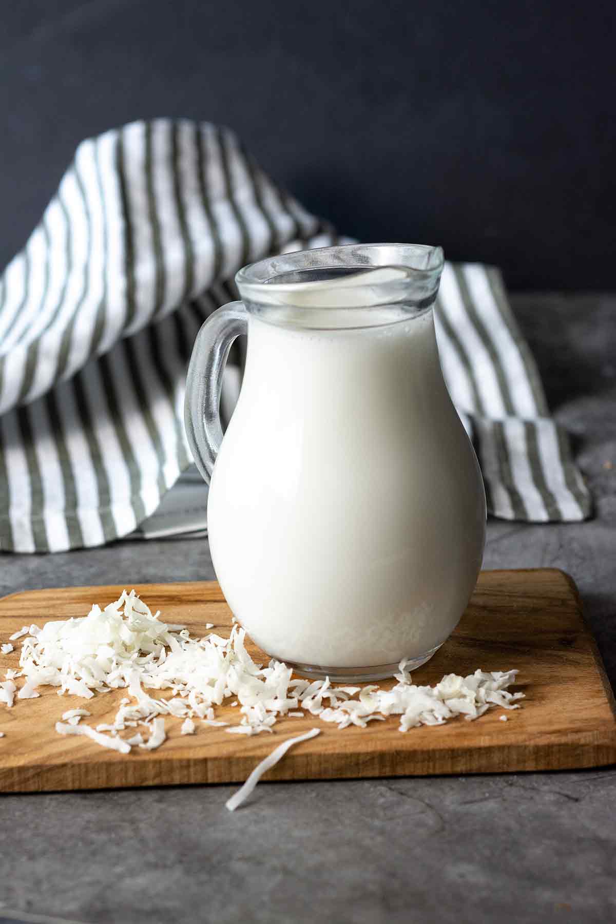 Coconut milk made from coconut flakes in a glass jar on a cutting board with coconut flakes and black and white stripe napkin in the background