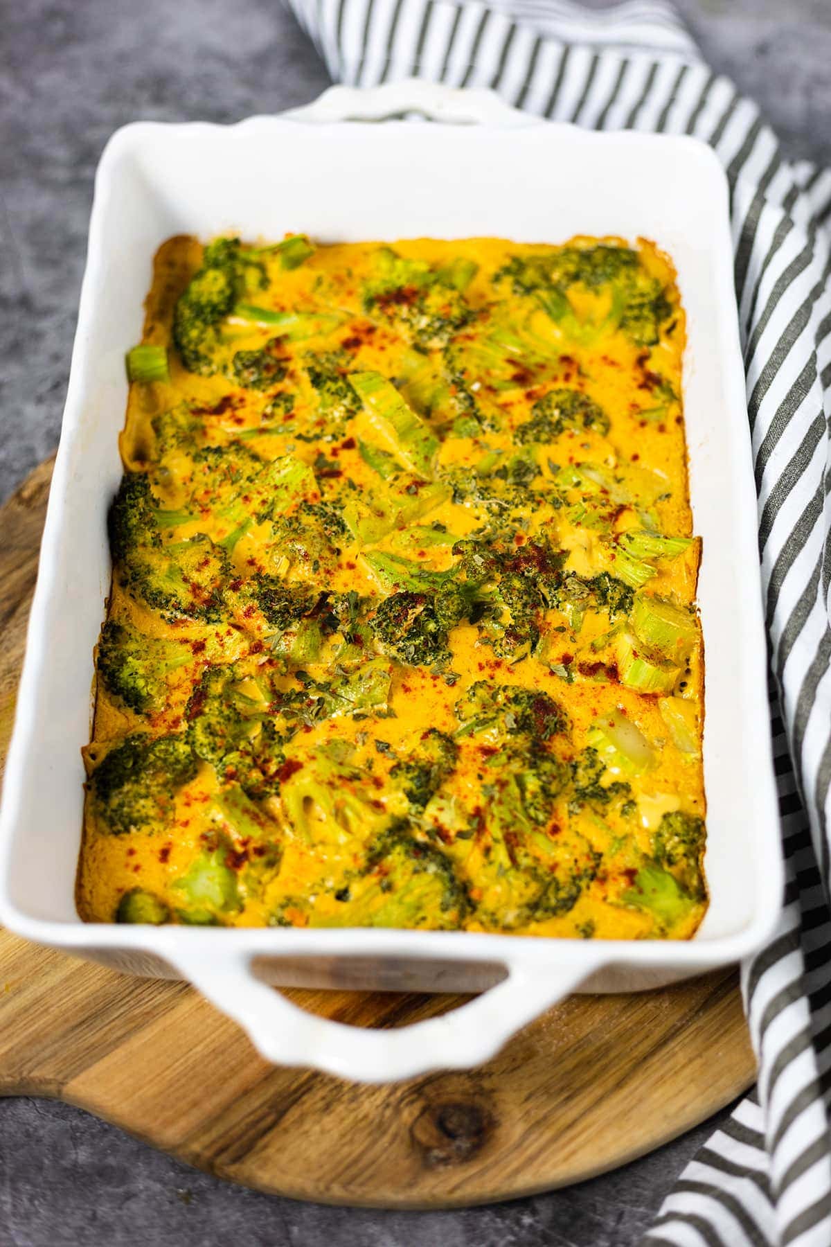 Broccoli Cheese Casserole in a white casserole dish on a cutting board with a black and white strip napkin