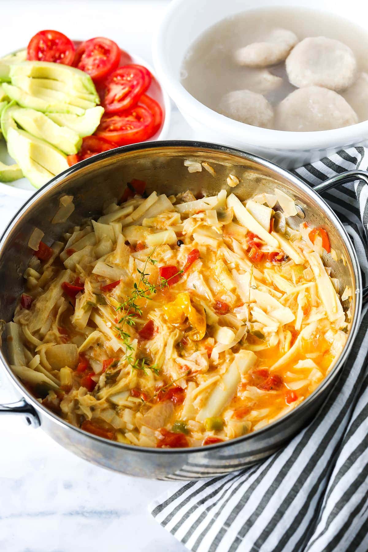 overlay vegan saltfish in a saucepan with dumplings in a white bowl, avocado and tomato slices on a white plate on a white background