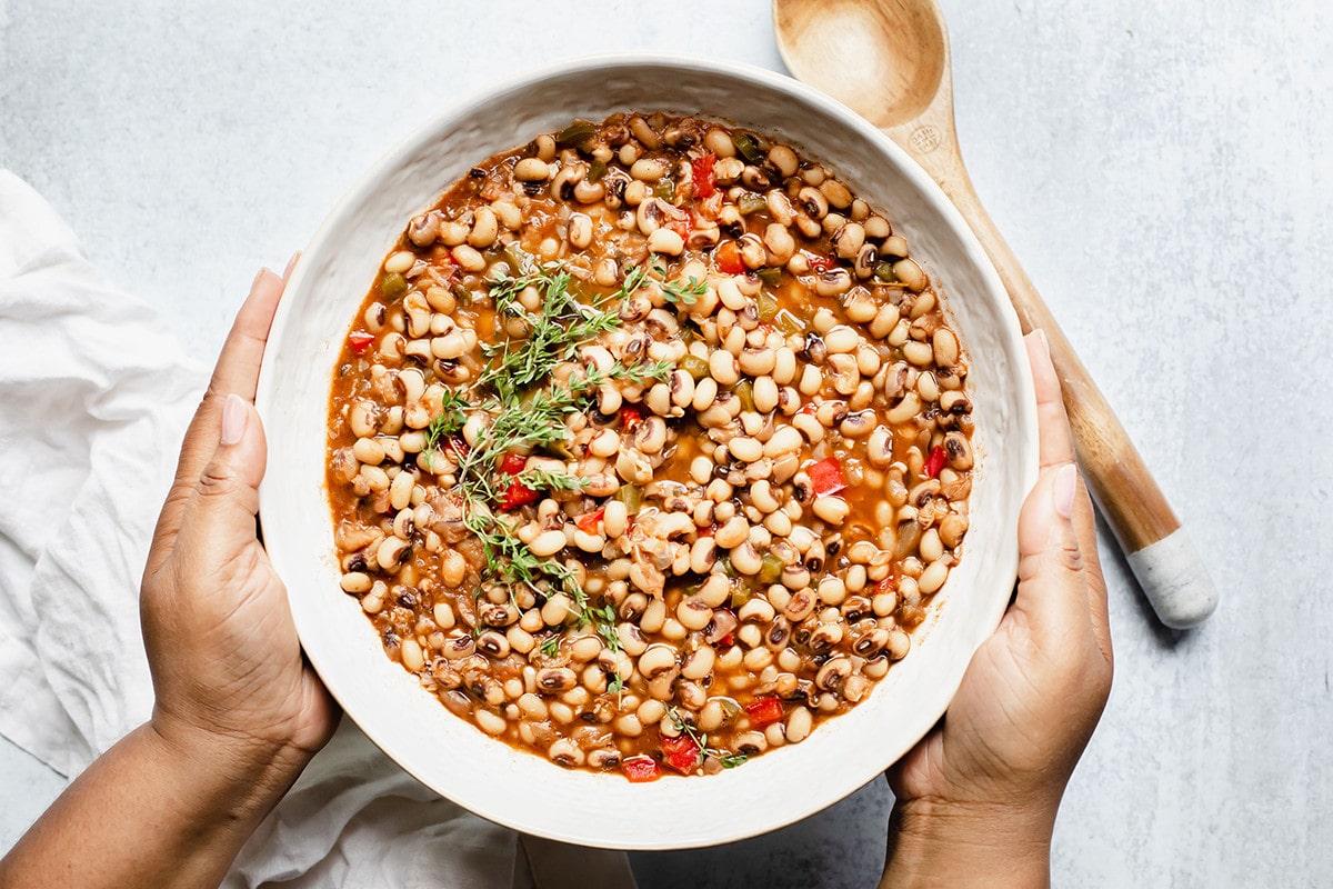 overlay southern black-eyed peas in a white bowl with a wooden spoon, being held with hands on a white background