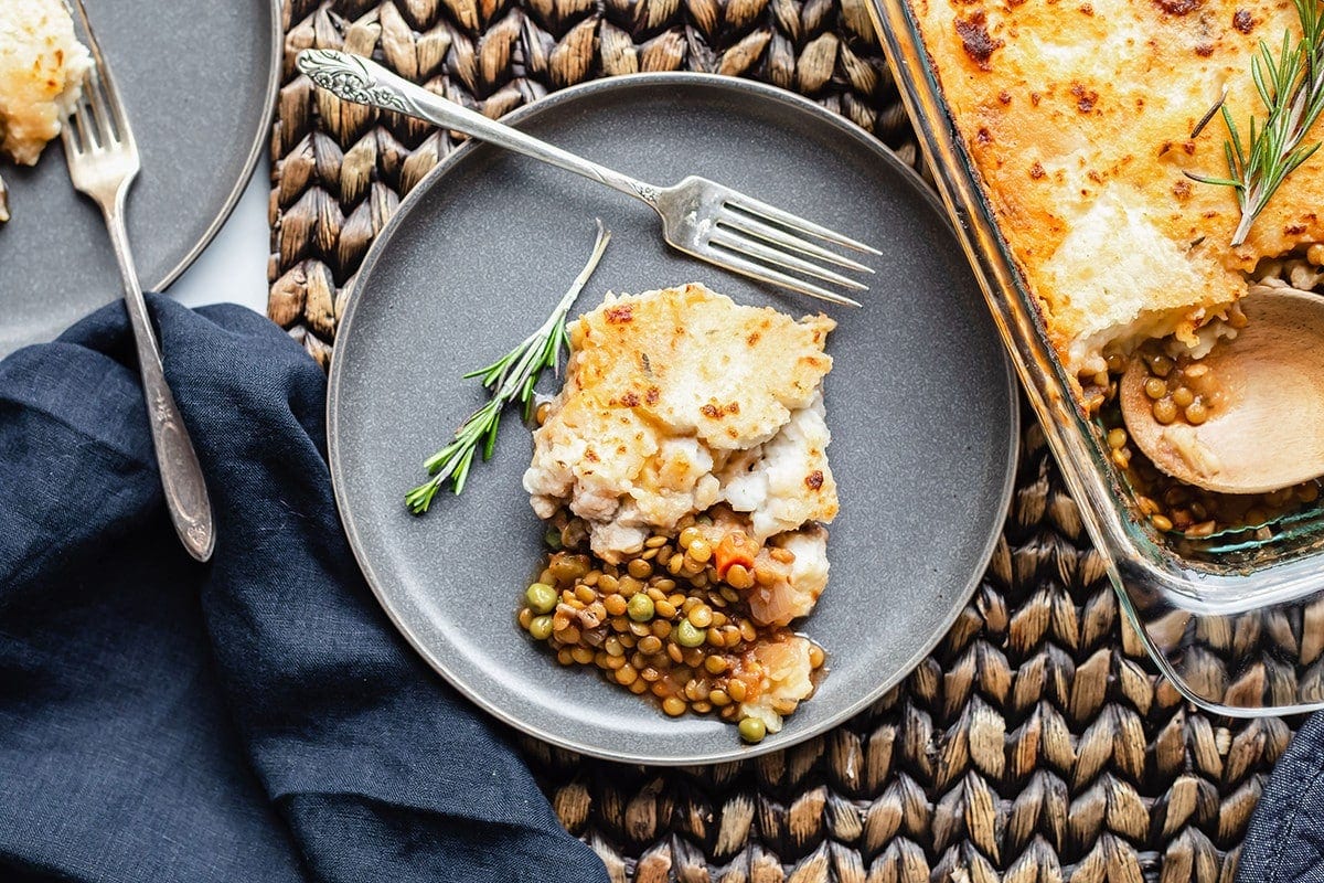 Overlay serving of lentil shepherd's pie on a grey plate with a fork and garnished with rosemary sprig with a blue napkin on a brown background