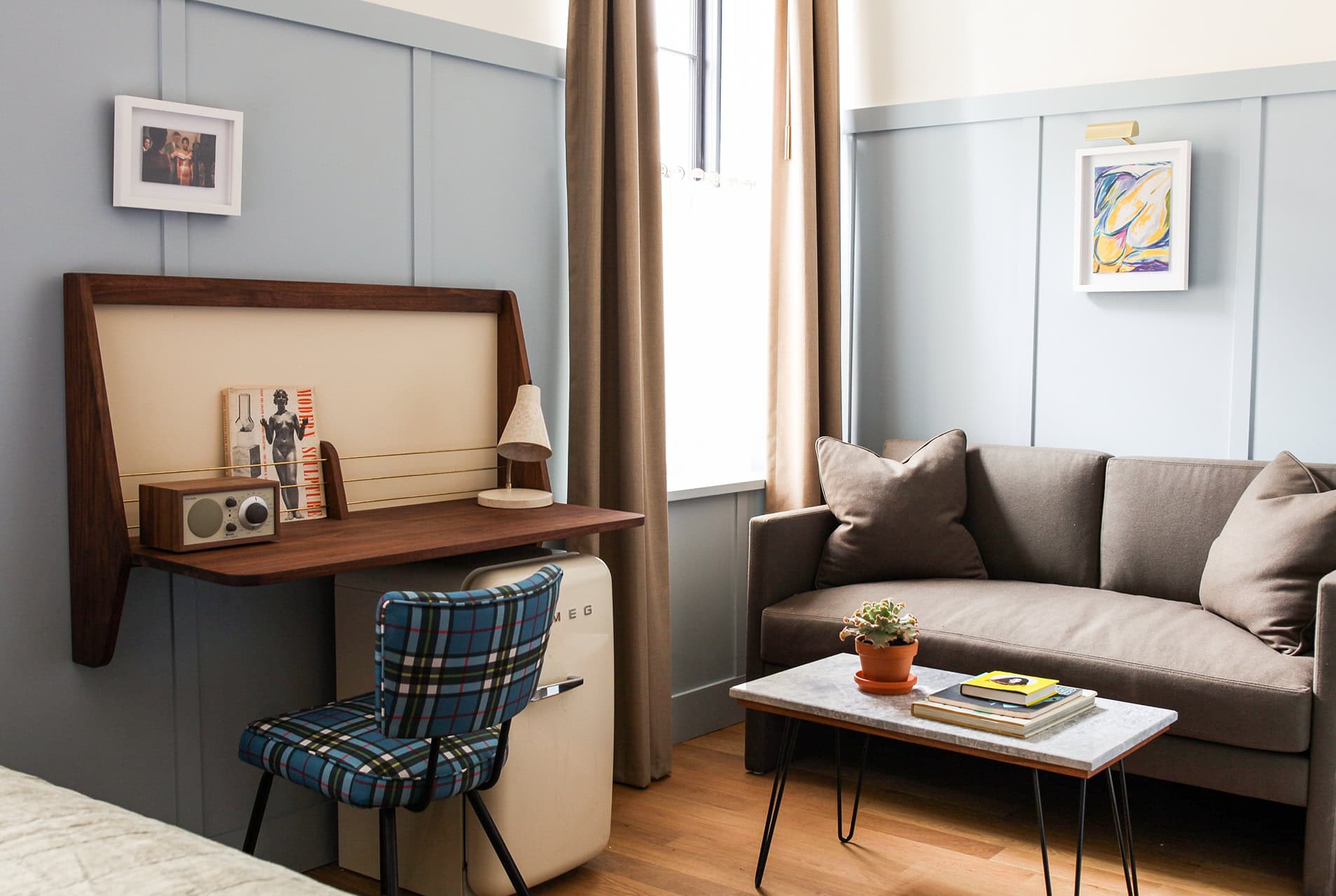 A compact but practical writing nook with sleek built-in MCM desk, cream Smeg mini fridge, next to a plush grey sofa in the Double Queen Guest Room at Hotel Kinsley's 301 Wall Street Location. Small accents and details, like the potted succulent, coffee table books and framed artworks make for an eclectic yet minimalist look.