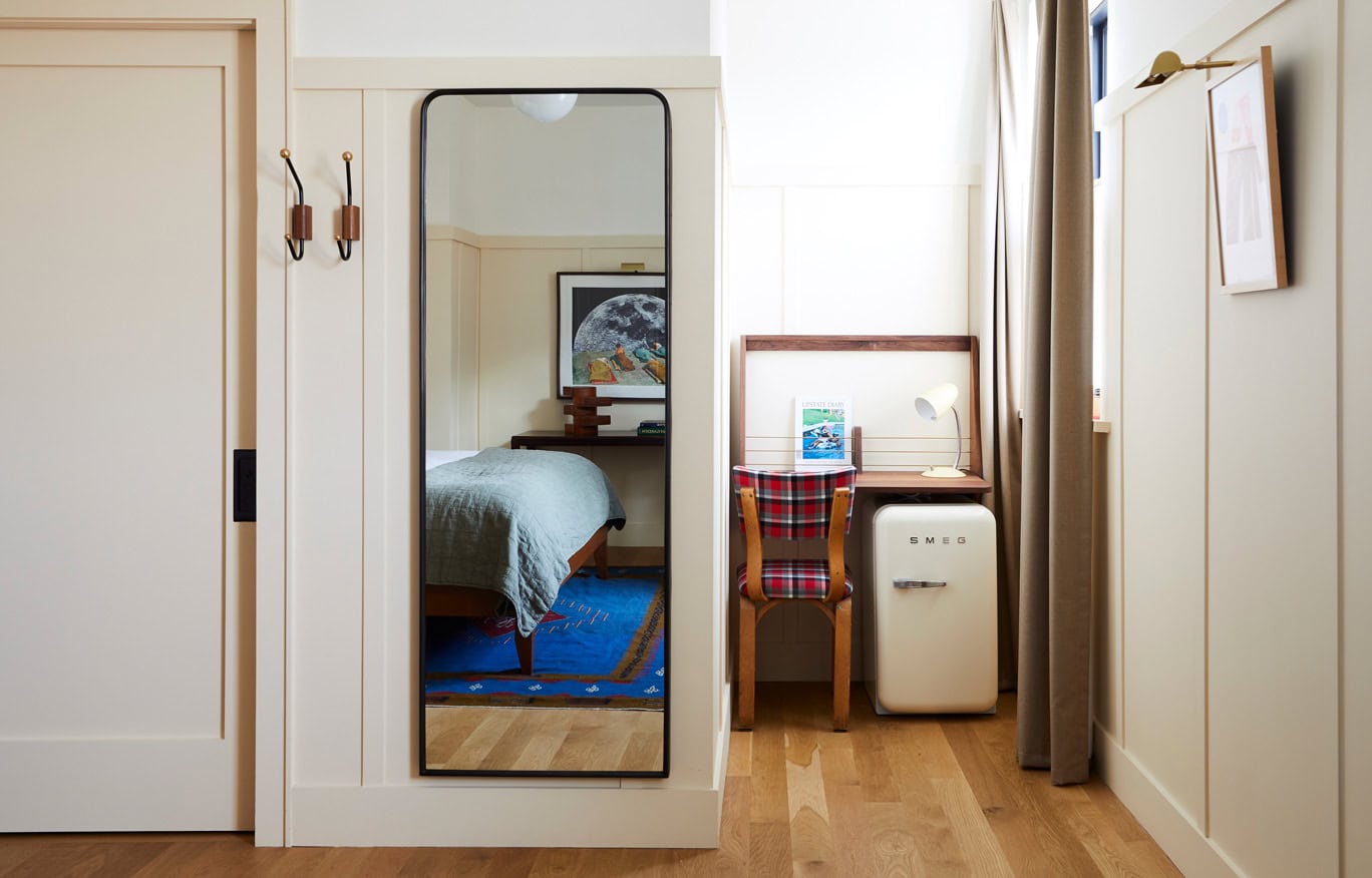 A compact but practical writing nook with cream Smeg mini fridge in a Standard King Guest Room at Hotel Kinsley's 301 Wall Street Location. A floor length modern mirror also shows a glimpse of the heavenly king size bed with luxury linens.