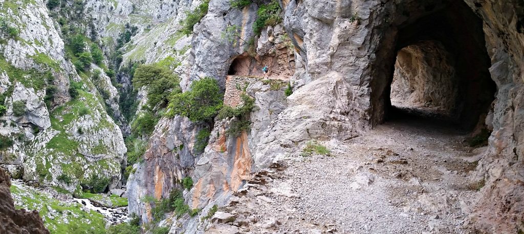Trail goes through rustic tunnel carved into rocky cliff in foreground. In background a tiny hiker is seen on the same trail far above rushing creek/river below.