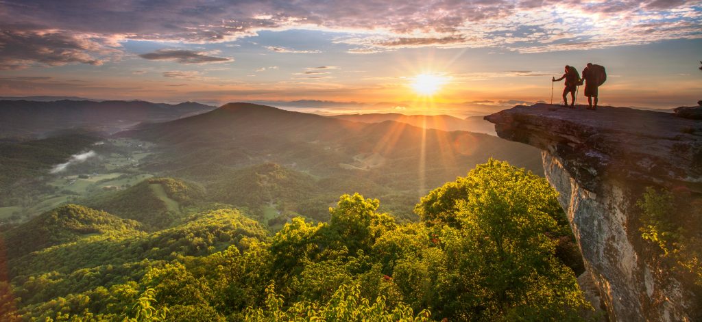 Two backpackers are silouetted by the sunrise as they stand on McAfee’s Knob on Appalachian Trail .