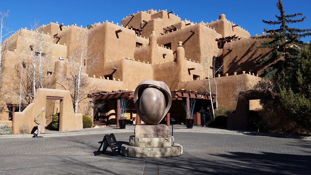 Exterior of six-story hotel designed after traditional pueblo with multiple flat roofs, log beams (vigas) sticking out of adobe-like walls. New Mexico blue sky behind.