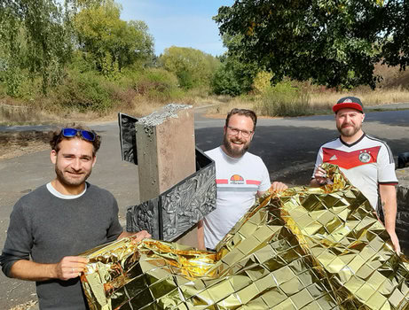 IFM-Vorstand vor dem Paul-Schneider-Denkmal in Pferdsfeld mit der goldenen Rettungsdecke als Zeichen der Initiative DIE VIELEN