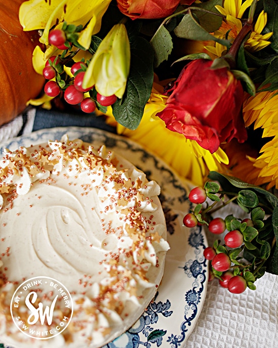 cheesecake with golden sprinkles on a blue and white plate surrounded by flowers and a pumpkin