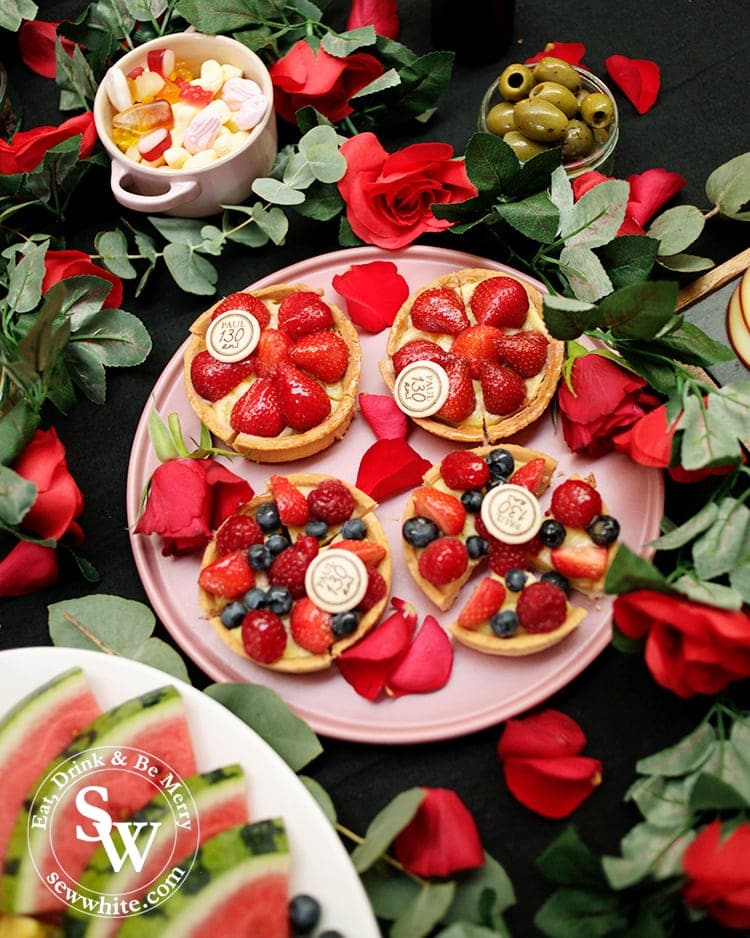 Beauty and the Beast Party table ideas. A dark tablecloth covered in Paul bakery strawberry tarts studded with berries. The plate wrapped in a rose garland and covered in rose petals.
