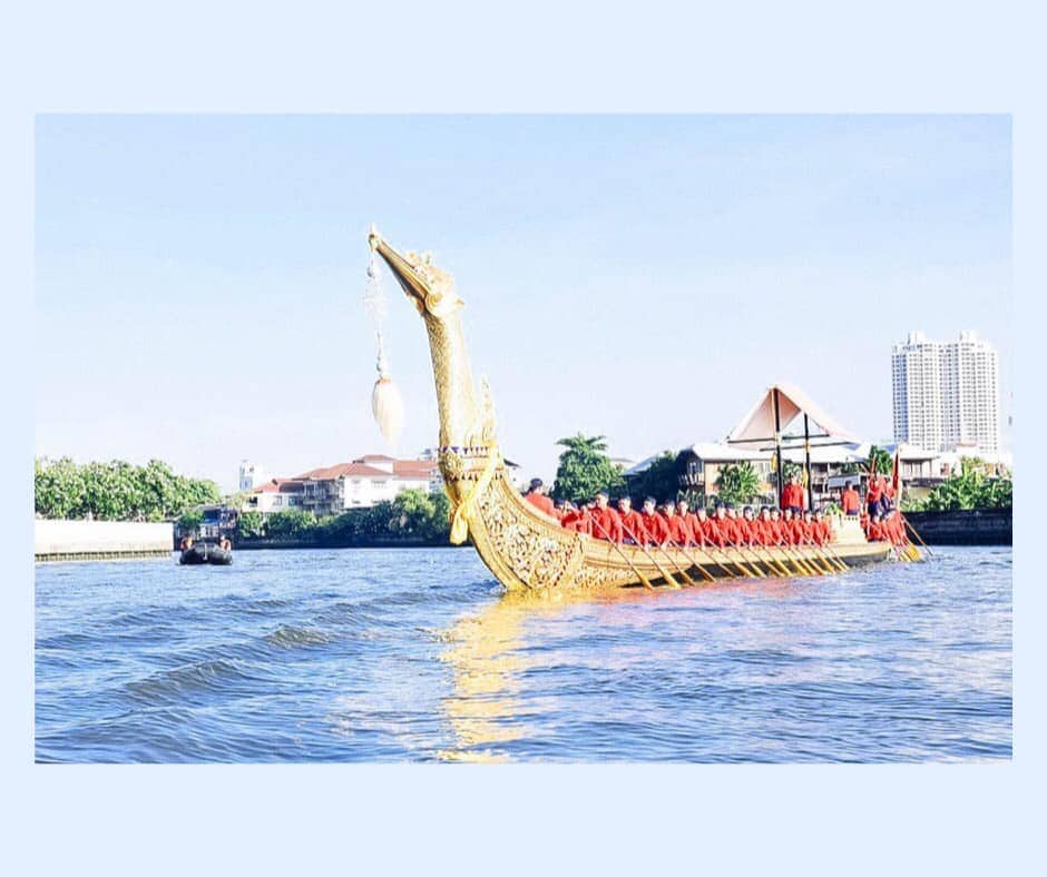 Golden swan is the bow of the gilded boat and points toward camera. ^0+ Thai Navy men dressed in traditional red regalia sit in the longboat. Blue water in foreground