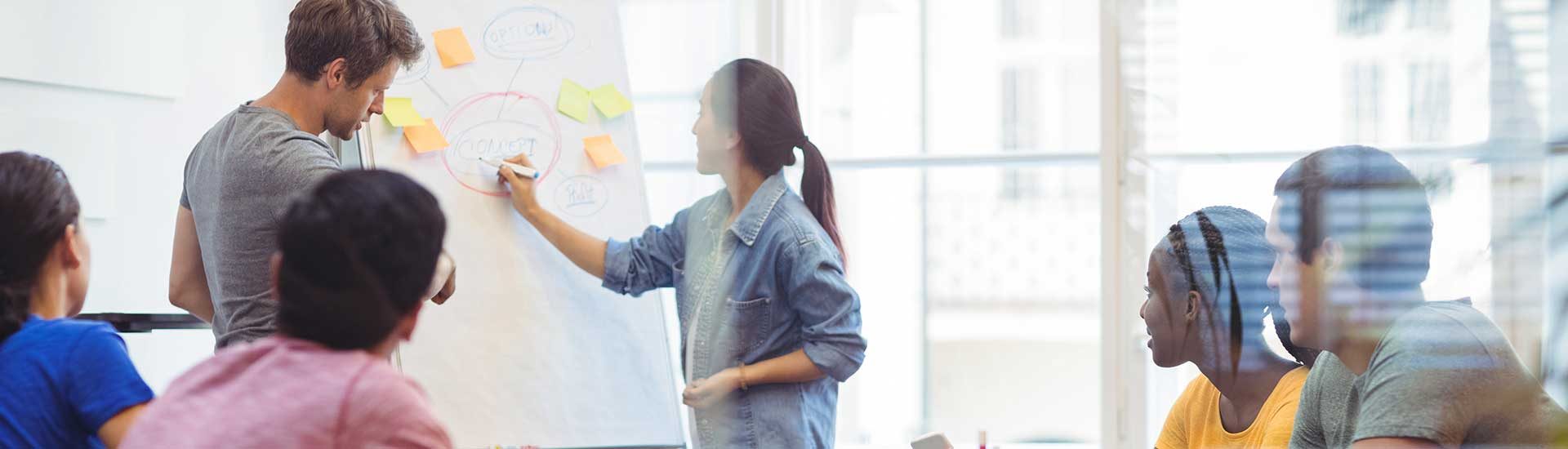 Jobnavigation – Über uns – Headerbild eine Frau schreibt etwas an ein Flipchart, eine Gruppe junger Menschen schaut zu