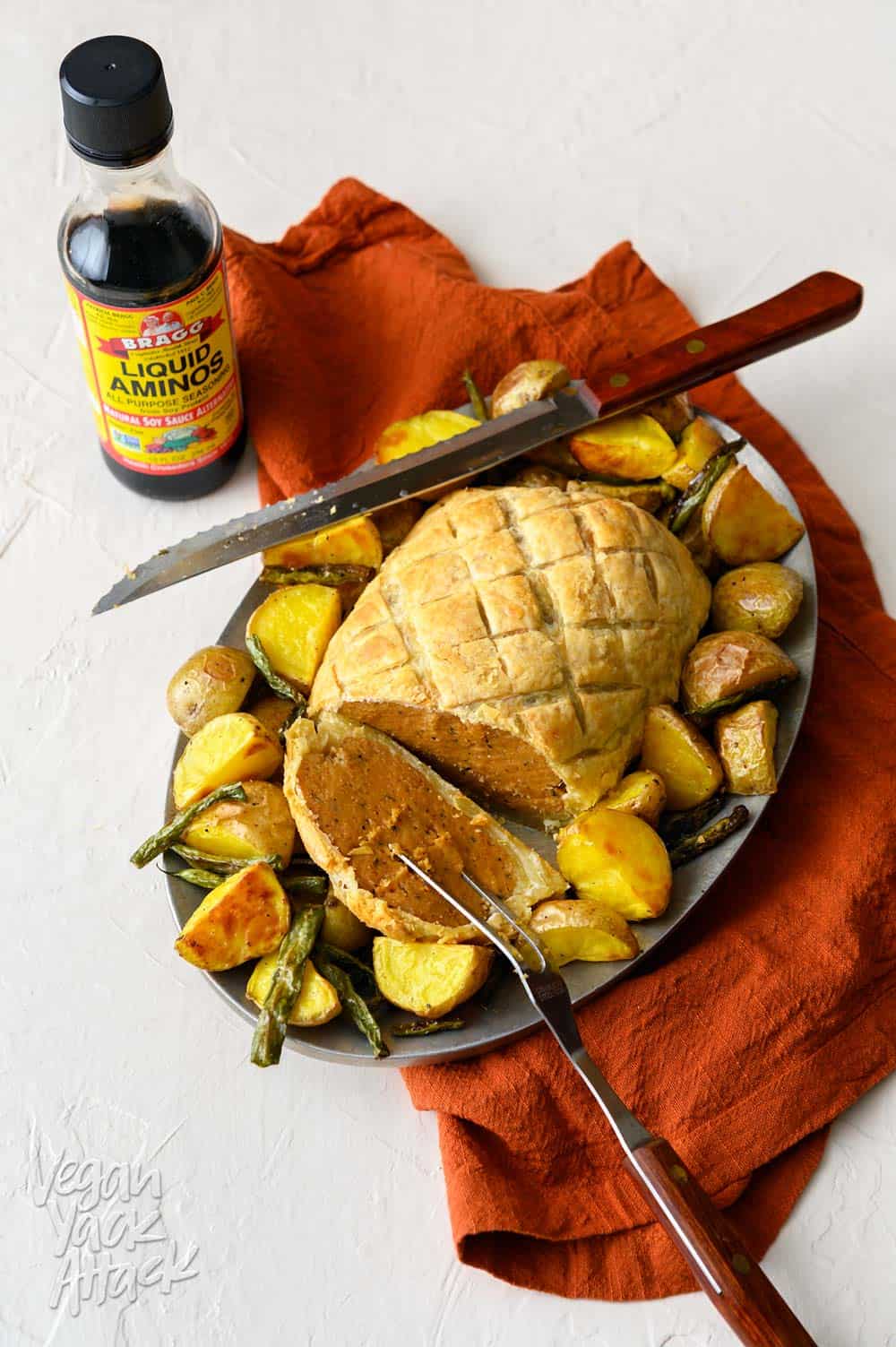 Seitan Wellington on a platter with roasted vegetables and rust-colored napkin, bottle of Bragg liquid aminos on the side