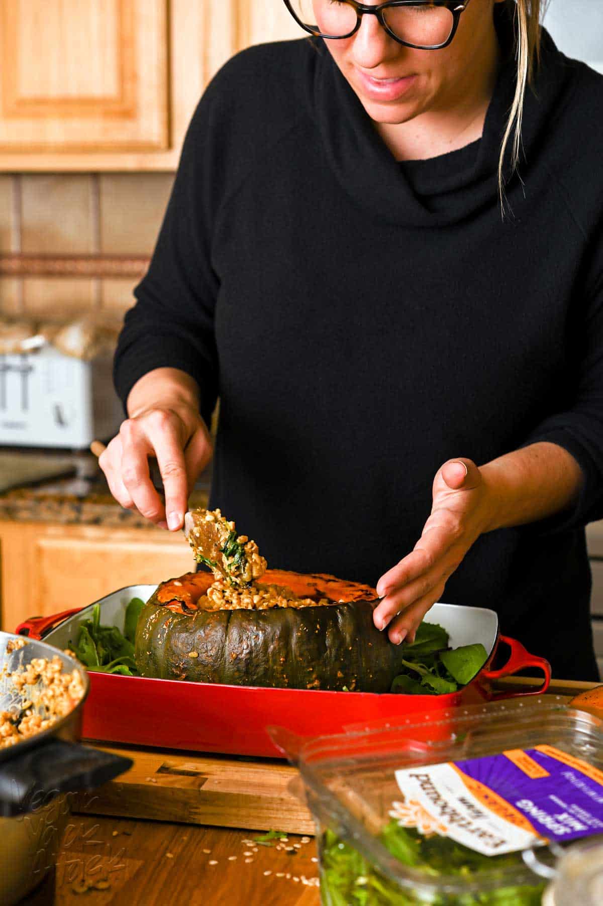 A woman in a black, long sleeve shirt, spooning rice stuffing into a large, roasted squash, that is in a red casserole dish.
