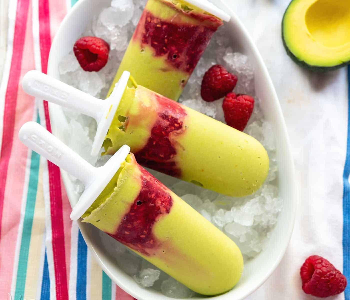 Green-hued avocado raspberry popsicles in a white dish on a striped linen