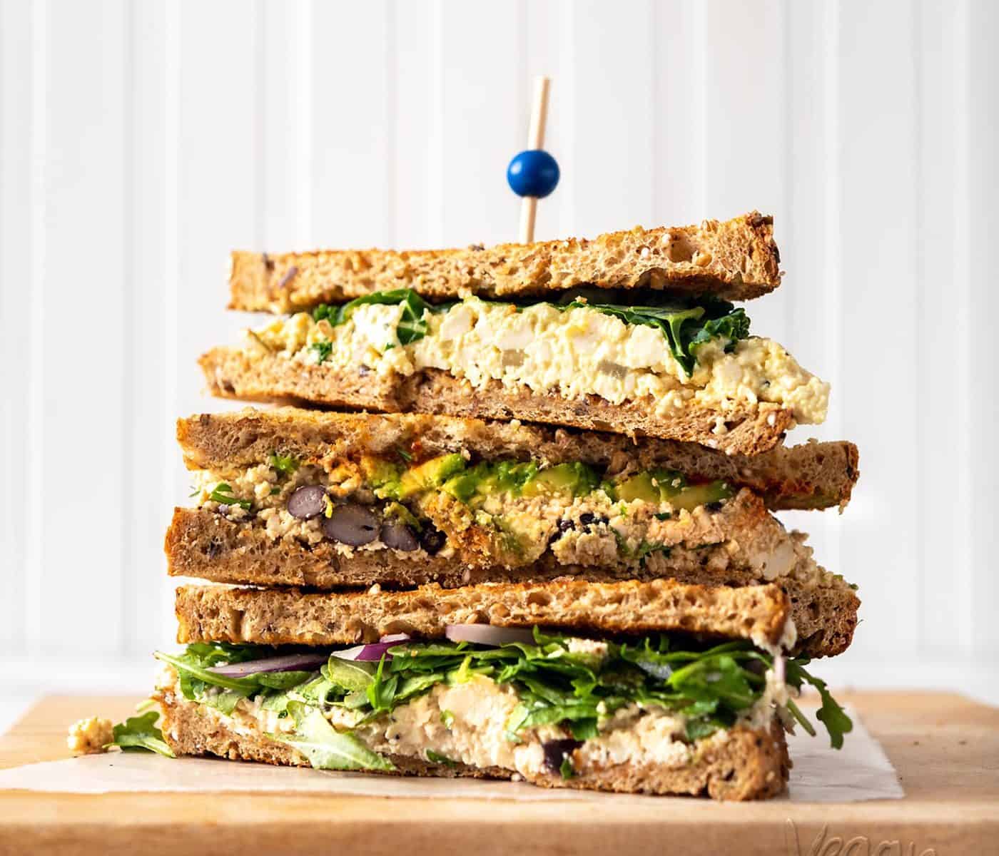Image of three tofu 'egg' salad sandwiches stacked on one another on a cutting board