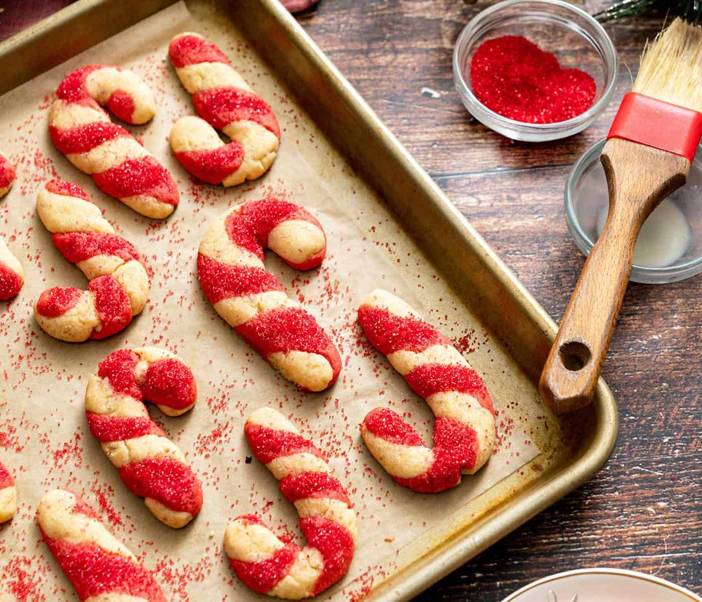 Candy cane-shaped cookies on a baking sheet next to colored sugar and pastry brush