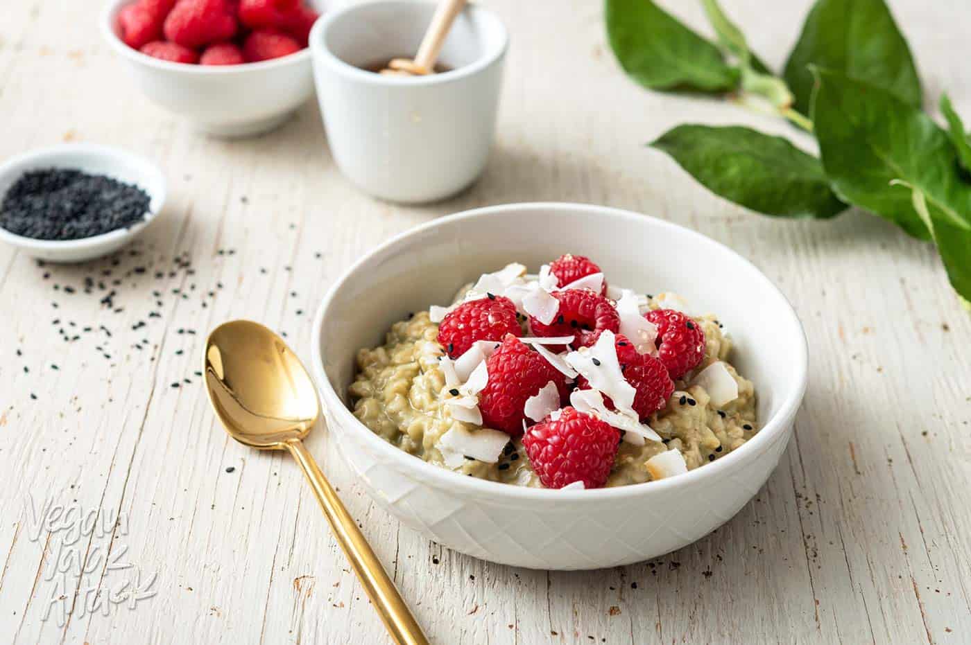 Image of white bowl with matcha oatmeal, topped with raspberries, on a white wood background, with leaves to the side