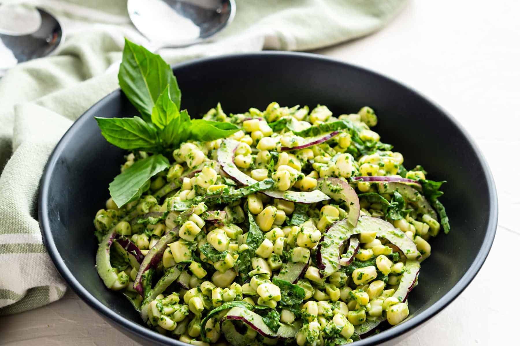 Large black bowl filled with Summer corn salad and basil pesto aioli with tongs and linen nearby