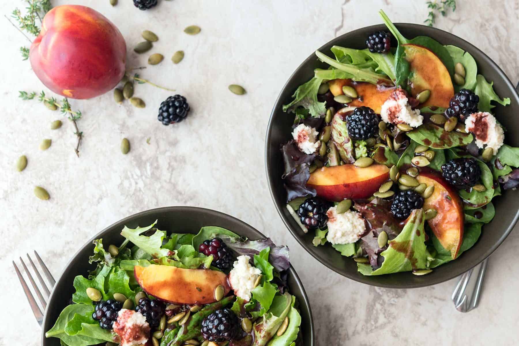 Three bowls of summery salad on a marble table top, next to seeds, a peach, and blackberries