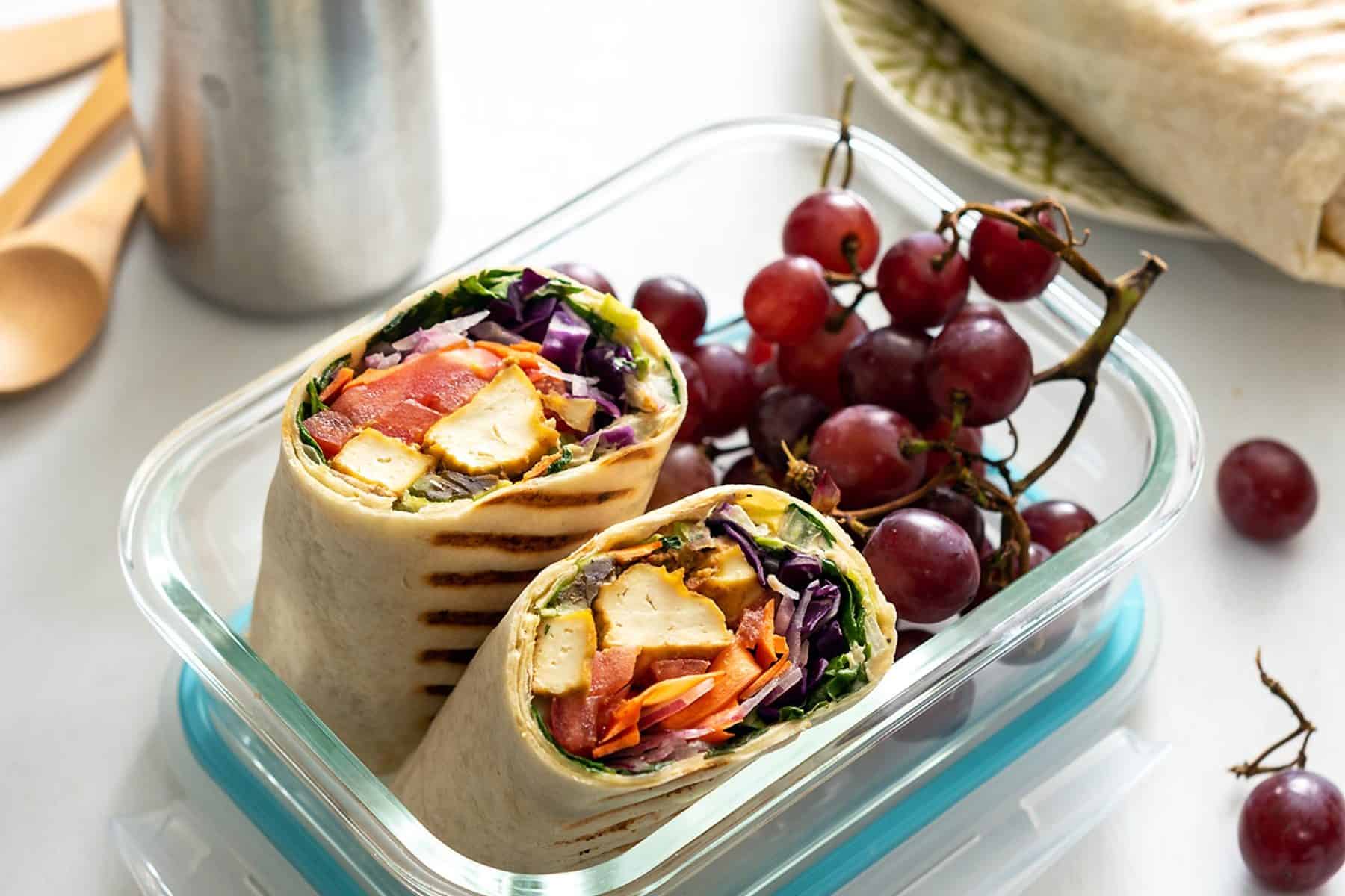 A chipotle tofu rainbow wrap cut in half in a lunch container with grapes
