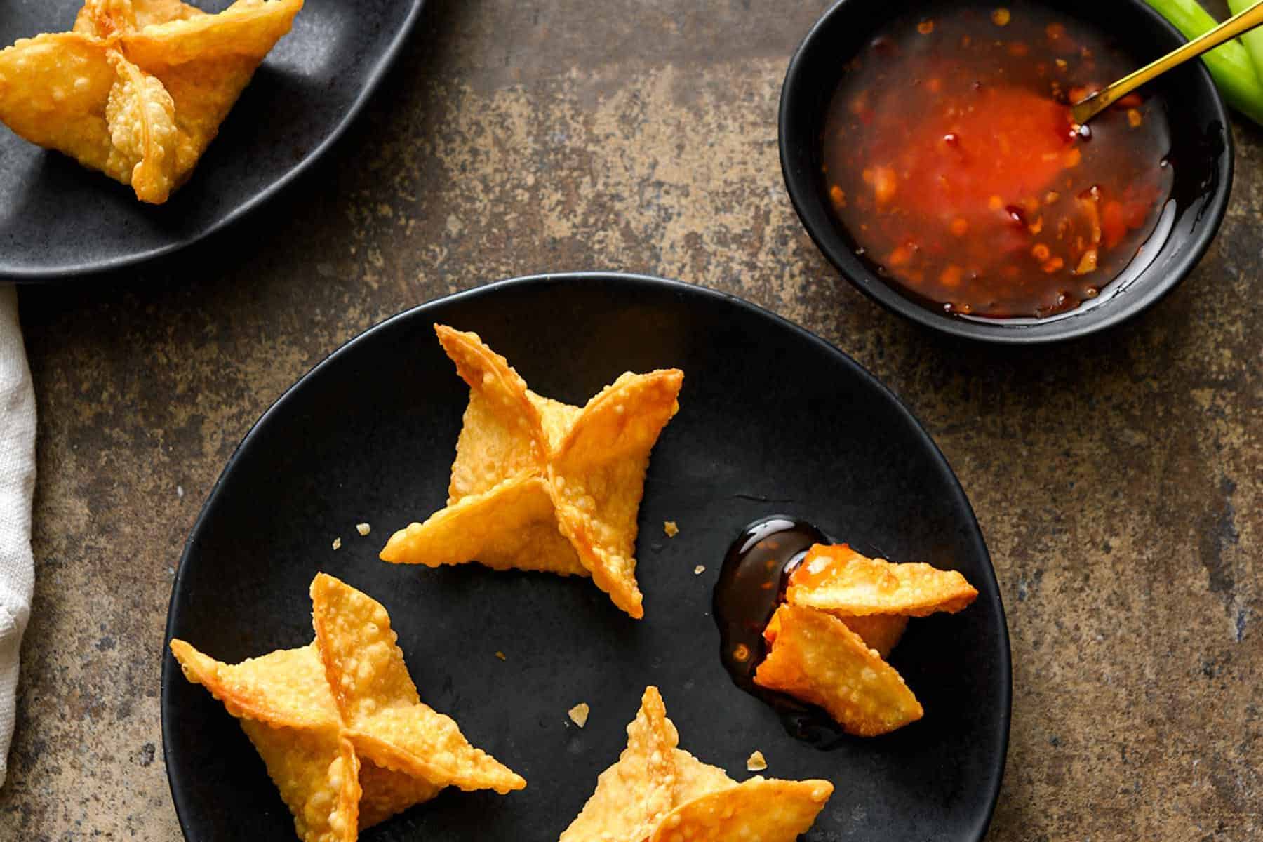Vegan Crab Rangoon on black plates with a side of sweet chili sauce, whole green onions, and a white linen on a great background