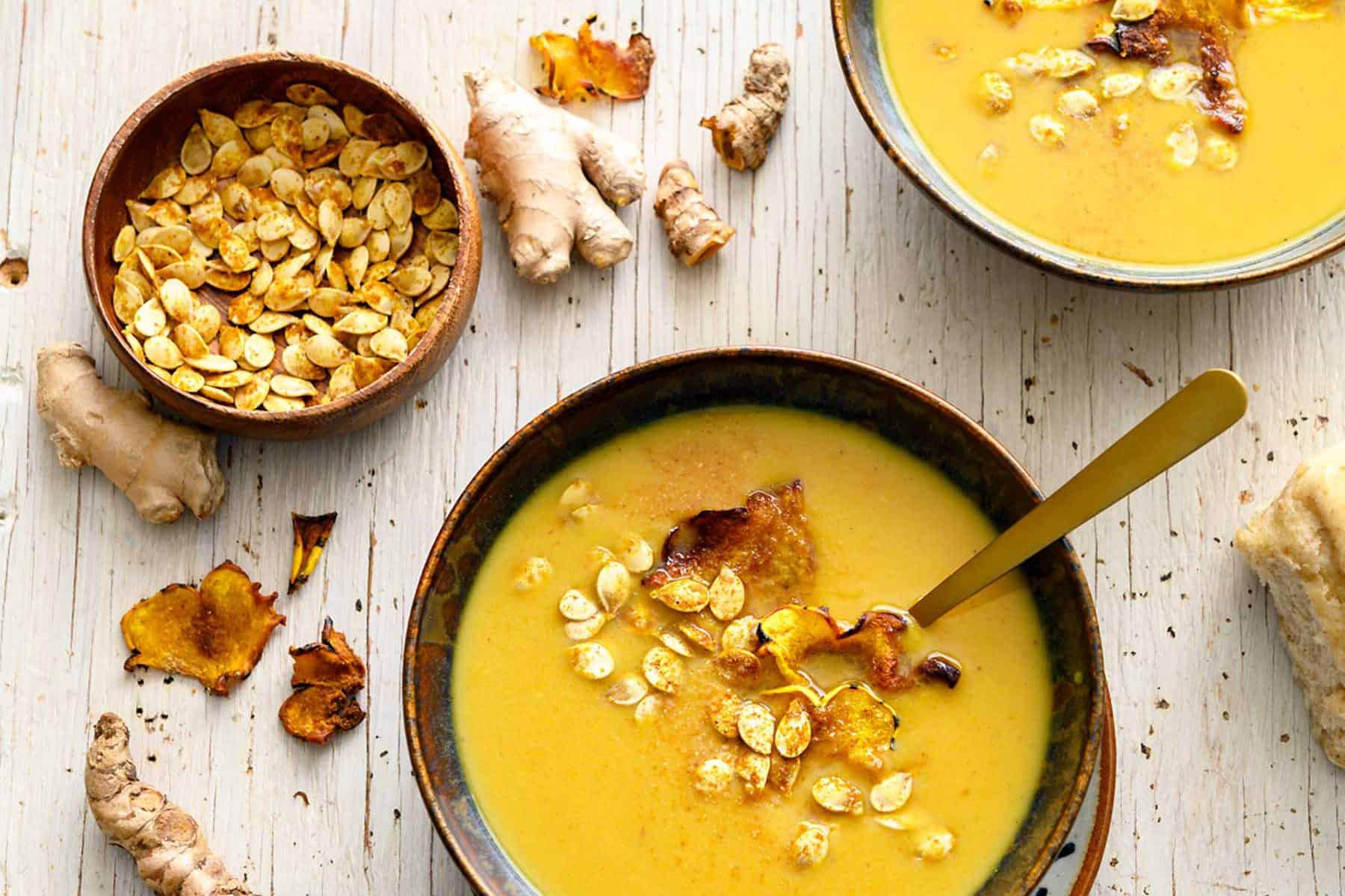 Two bowls of ginger turmeric squash soup with toasted seeds and a bread roll
